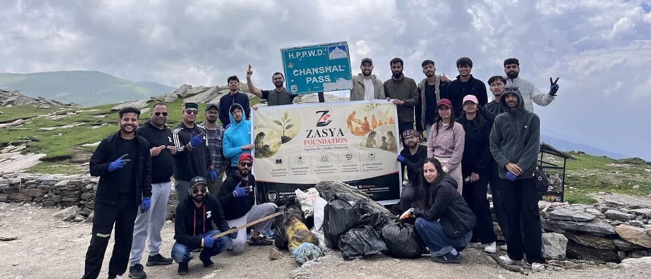 About twenty people in outdoor jackets stand on a grassy mountain pass holding a Zasya Foundation banner, beside a road sign reading H.P.P.W.D. Chanshal Pass, with filled refuse sacks gathered on the ground in front of them.