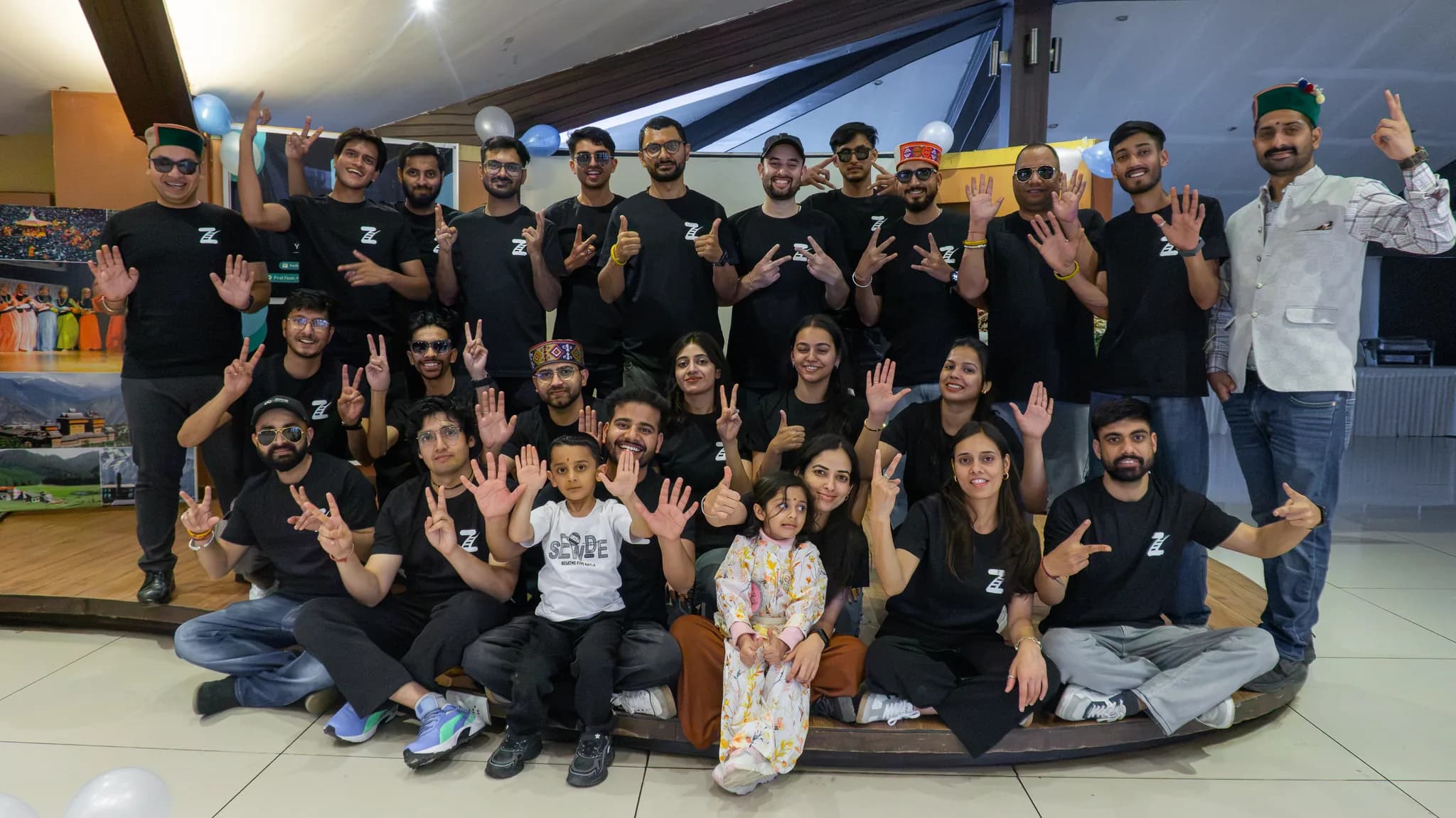 The Zasya team posed in three rows on a low stage at an indoor event, most in black t-shirts carrying the Zasya mark, with balloons behind them and two young children seated at the front.